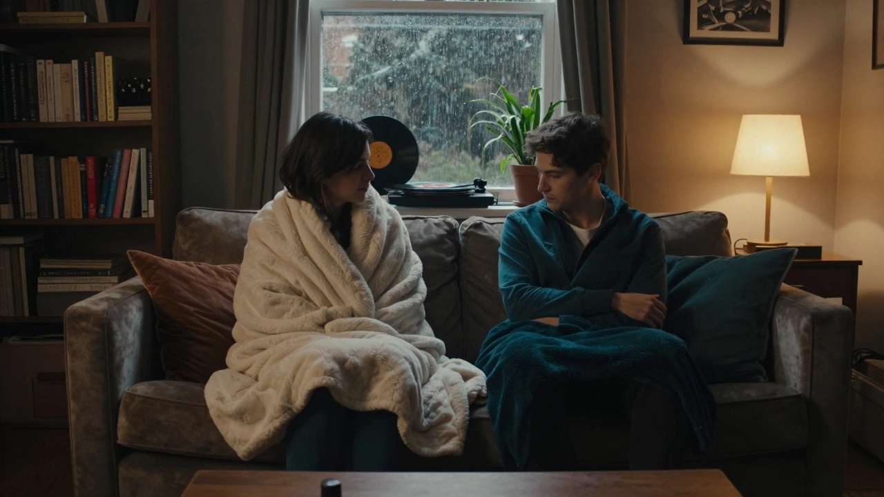Two individuals on a sofa in a quiet Haggerston flat, wrapped in blankets, sharing silence as rain falls outside.